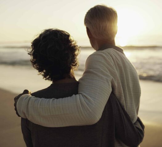 Två personer står på en strand och håller om varandra medan de ser ut över havet i solnedgången.