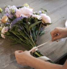 Händer knyter ett band runt en bukett med blandade blommor på ett bord.