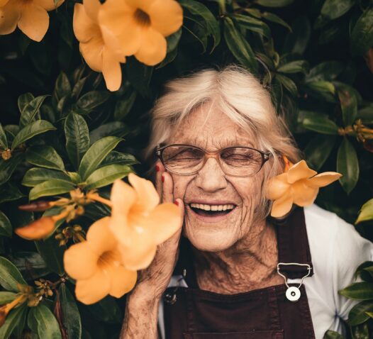 En äldre kvinna ler omgiven av gröna blad och orange blommor.